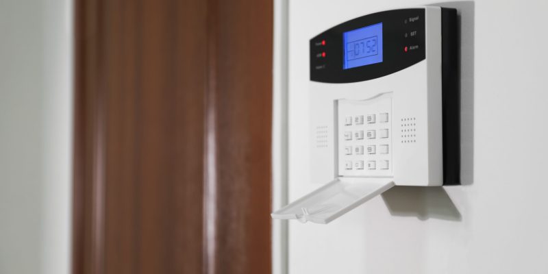 Home security alarm system on white wall near door, space for text